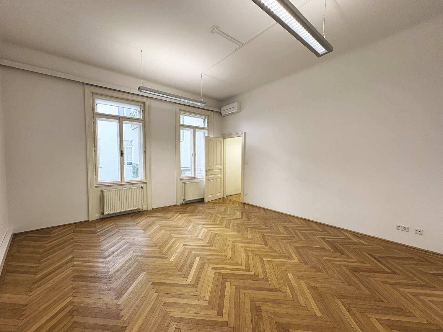 Geräumiges Zimmer mit Fischgrätparkett, hohen Fenstern und Klimaanlage, bereit für individuelle Gestaltung.