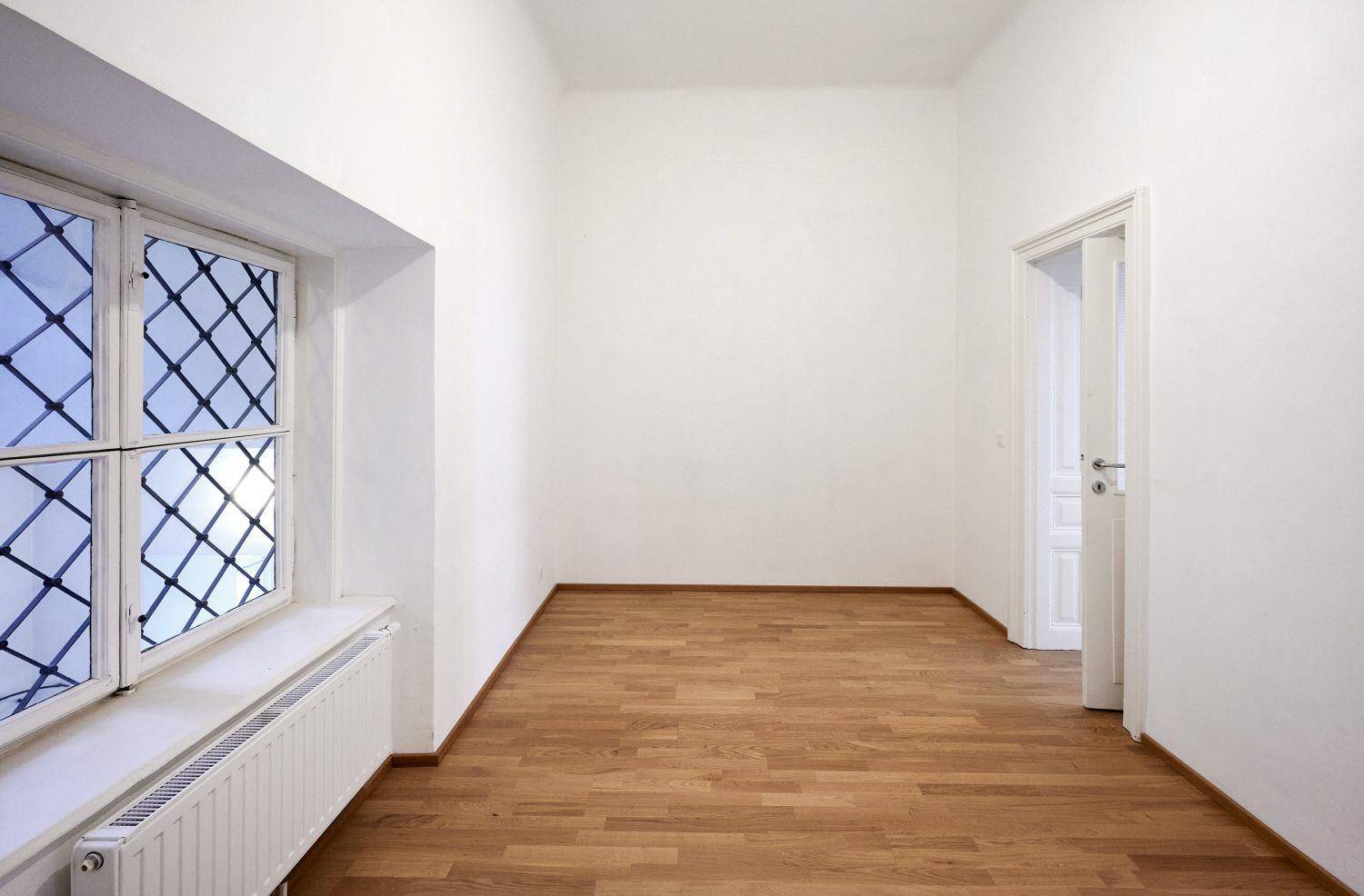 Leerer Raum mit Parkettboden, weißen Wänden und einem Fenster mit Gitter.