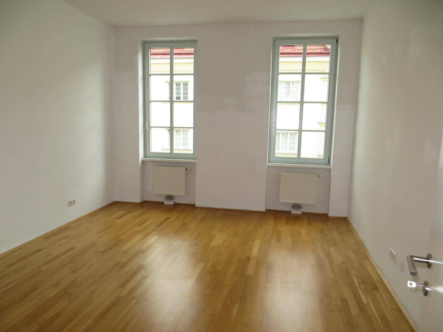 Helles, leeres Schlafzimmer mit Parkettboden und zwei hohen Fenstern, die den Raum erhellen.