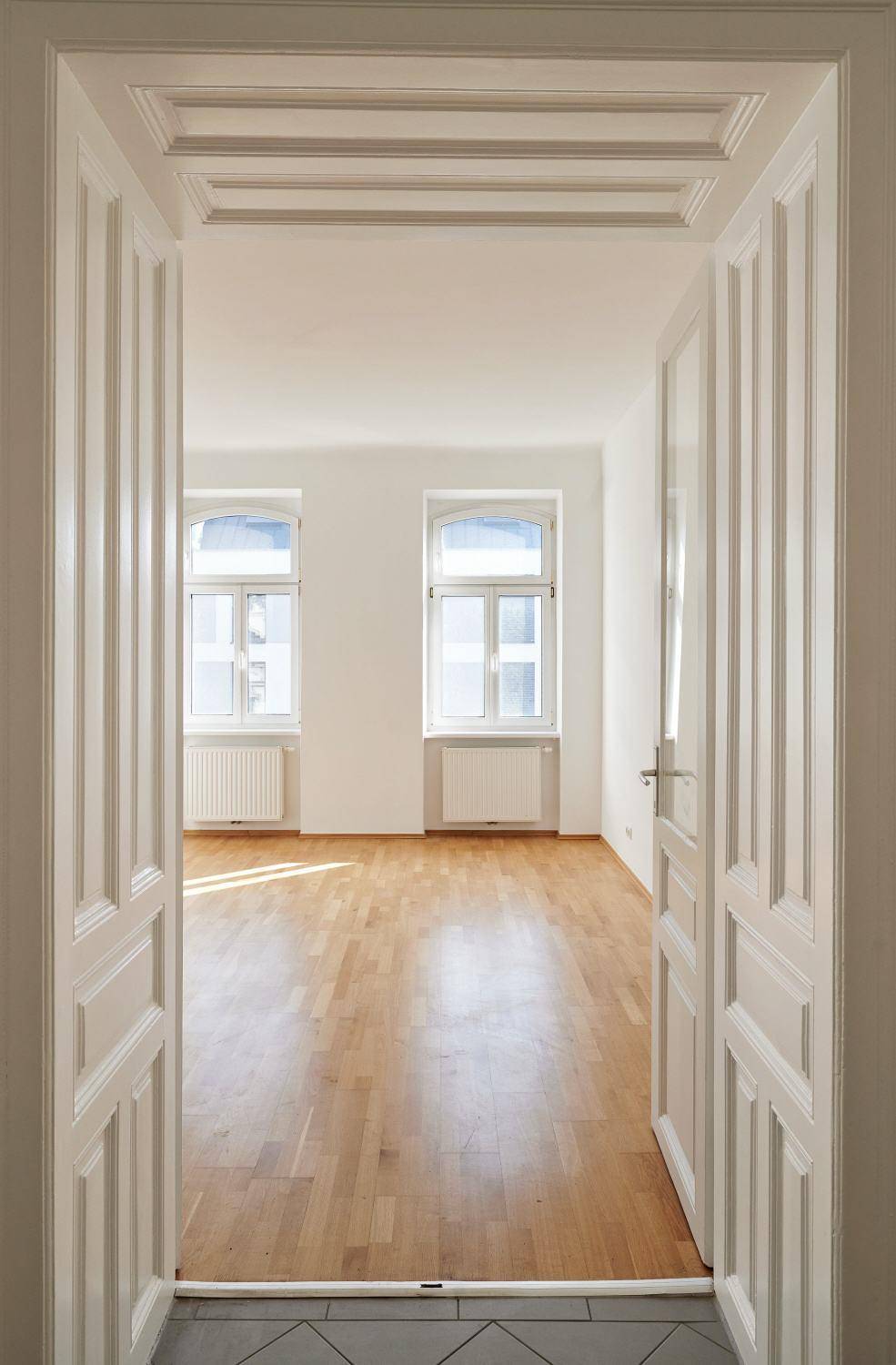 Blick durch eine klassische Flügeltür in ein helles Zimmer mit Parkettboden und zwei Fenstern.