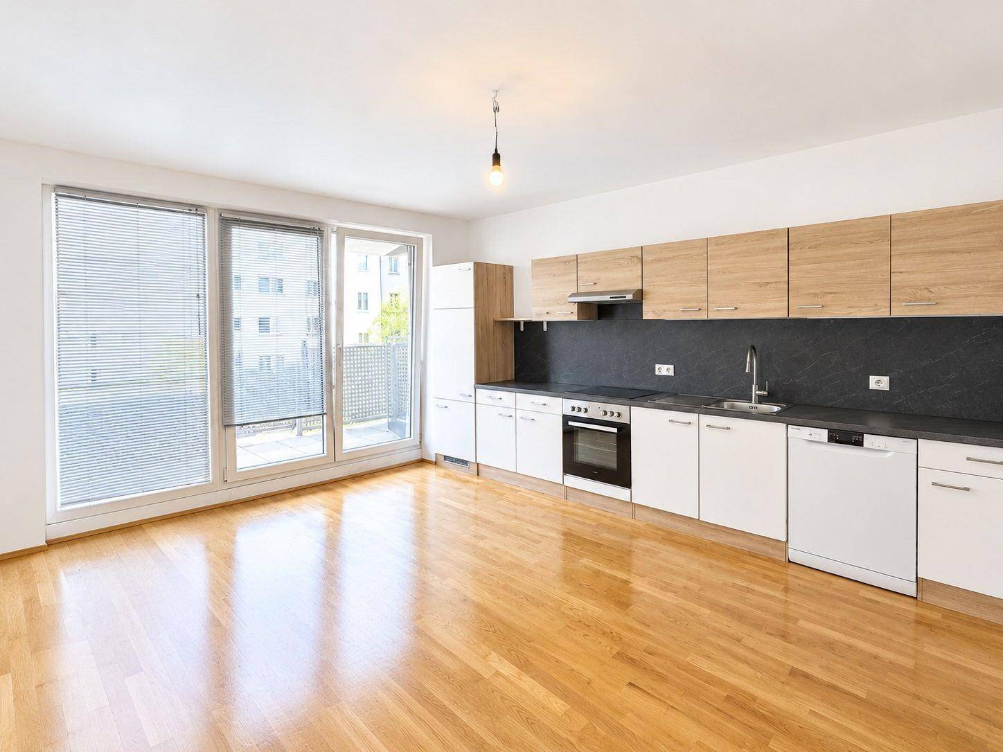 Heller offener Wohnbereich mit integrierter Küche, Parkettboden und Zugang zum Balkon durch große Fenster.