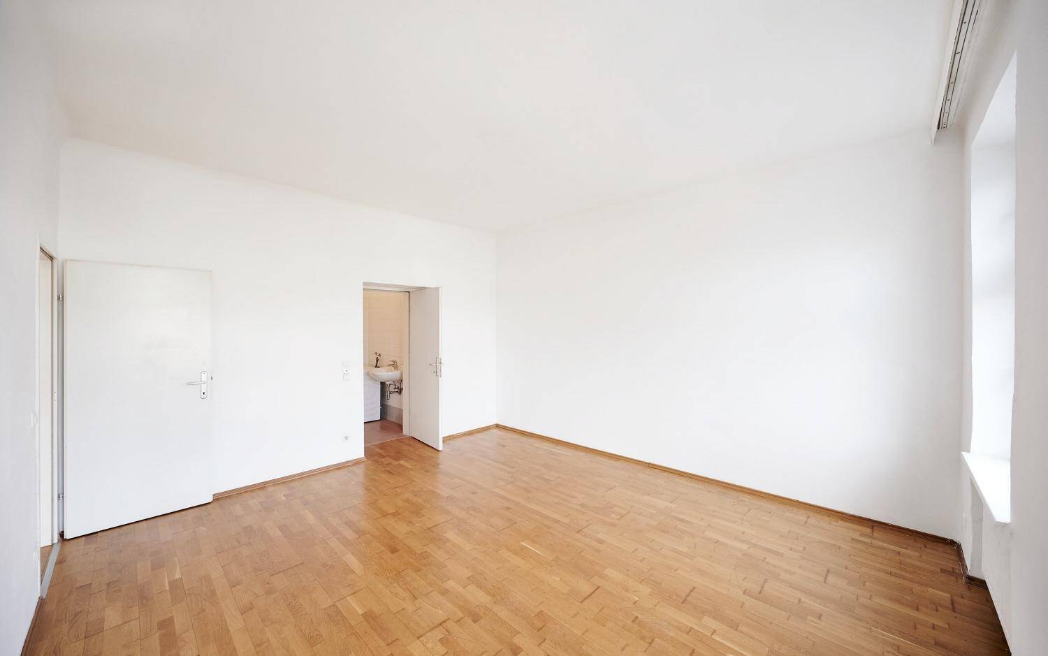 Heller Raum mit Holzboden und einer Tür, die zu einem angrenzenden Badezimmer führt.