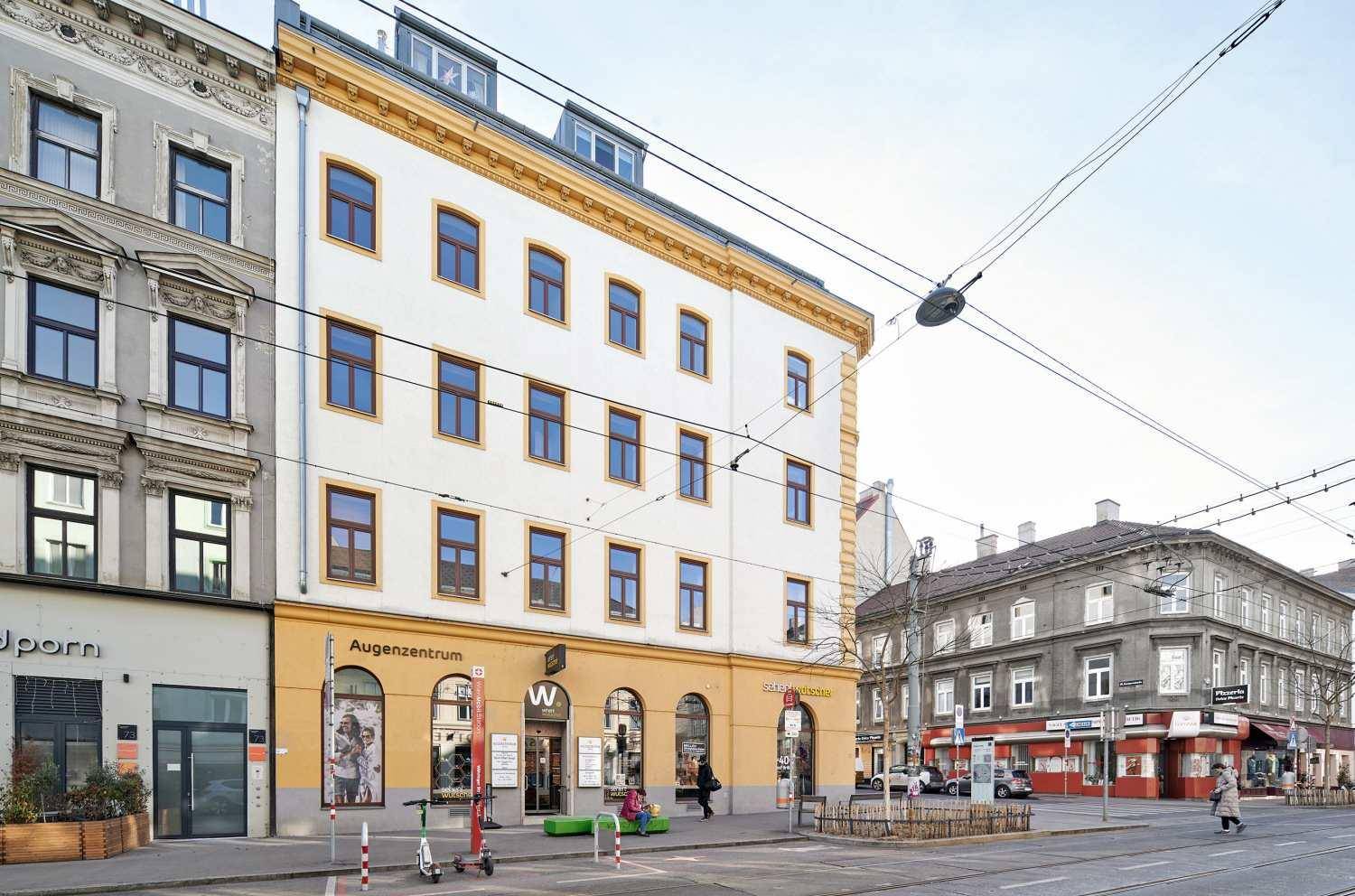 Eckgebäude mit Geschäften im Erdgeschoss an einer städtischen Straße mit Passanten und Verkehr.