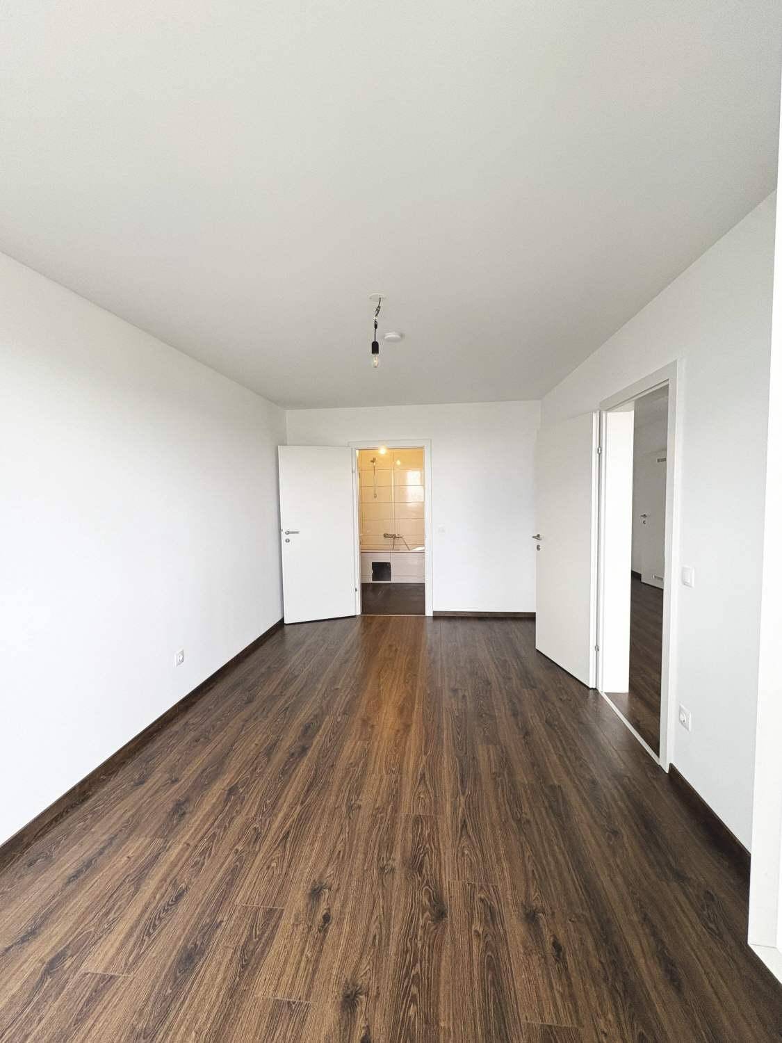 Heller Raum mit dunklem Laminatboden und weißen Wänden, der zu einem Badezimmer führt.