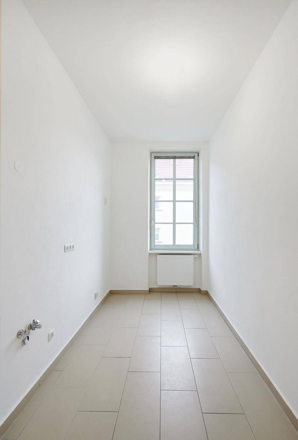 Schmaler Raum mit Fliesenboden und Fenster, vorbereitet für eine mögliche Küchen- oder Nutzungsinstallation.