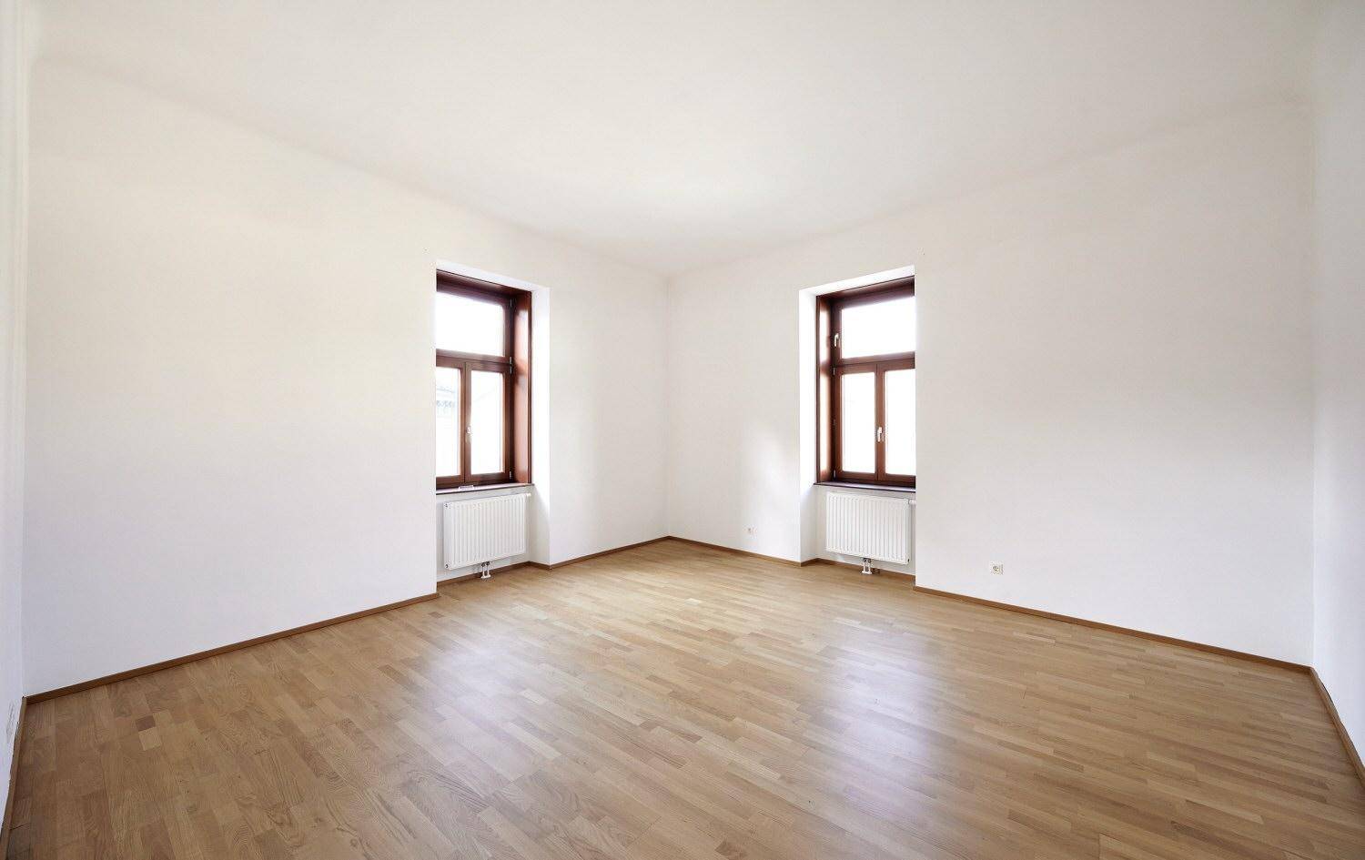 Leerer Raum mit Parkettboden, weißen Wänden und zwei Fenstern.