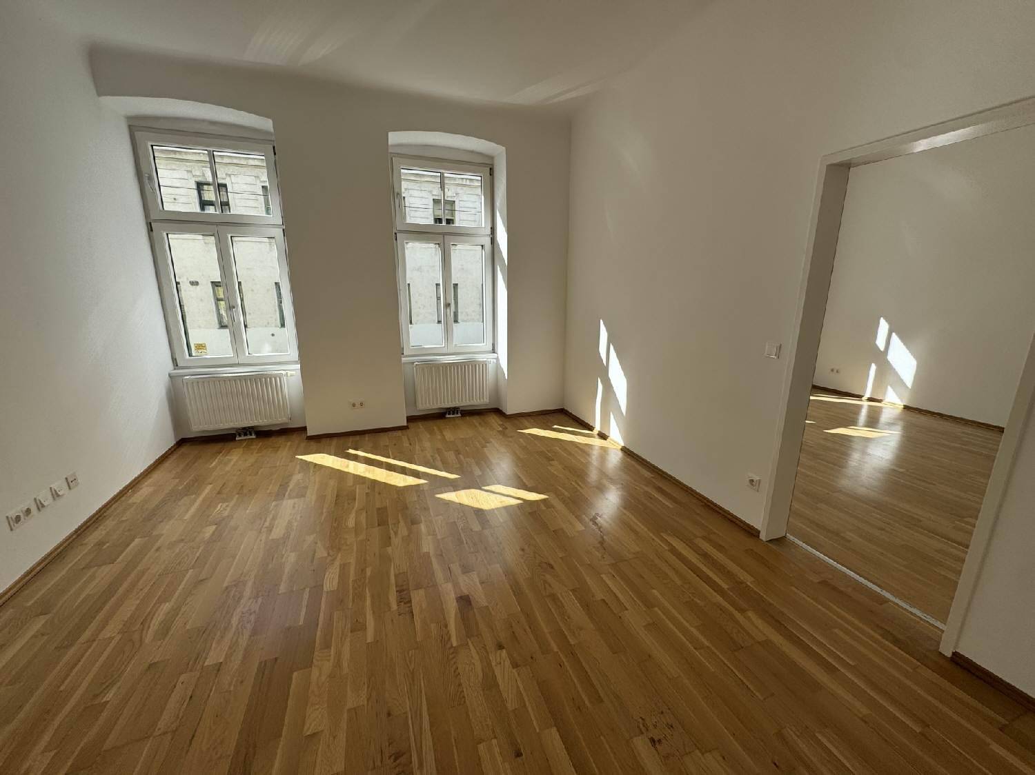 Lichtdurchflutetes Zimmer mit Holzboden und zwei hohen Fenstern, die den Raum erhellen.
