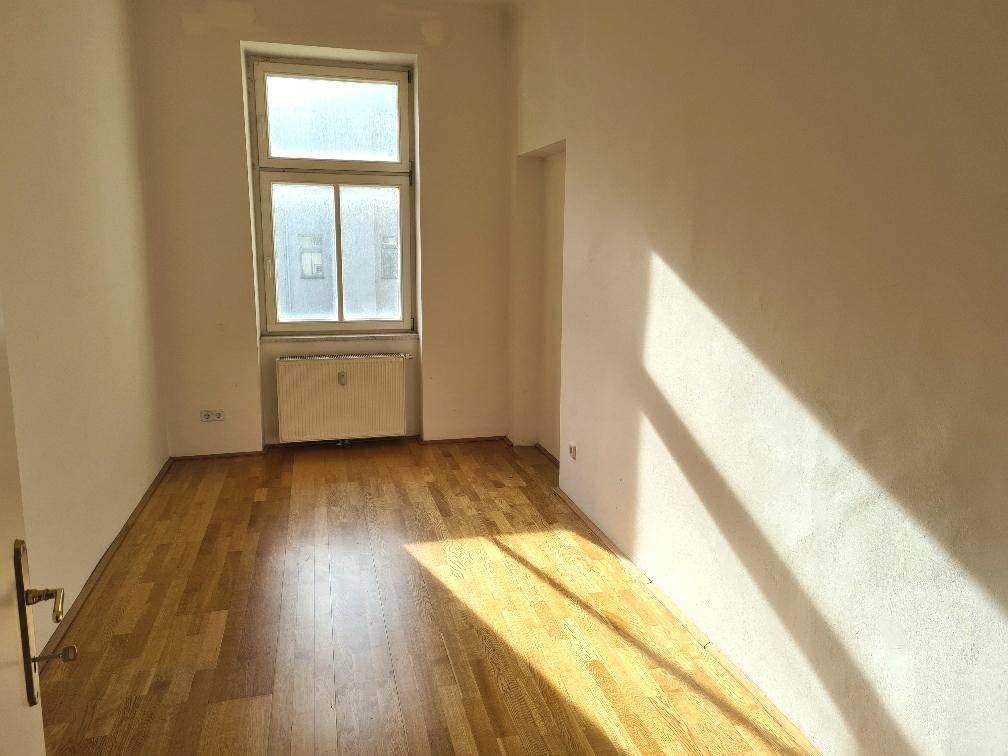 Heller, leerer Raum mit schönem Parkettboden und großem Fenster, durch das viel Sonnenlicht einfällt.