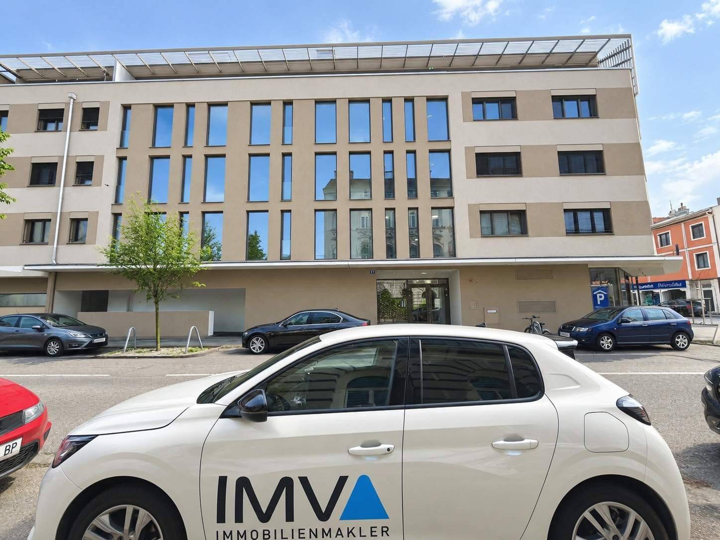 Moderne Gebäudefassade an einer Straße mit geparkten Autos und einem Immobilienmaklerfahrzeug.