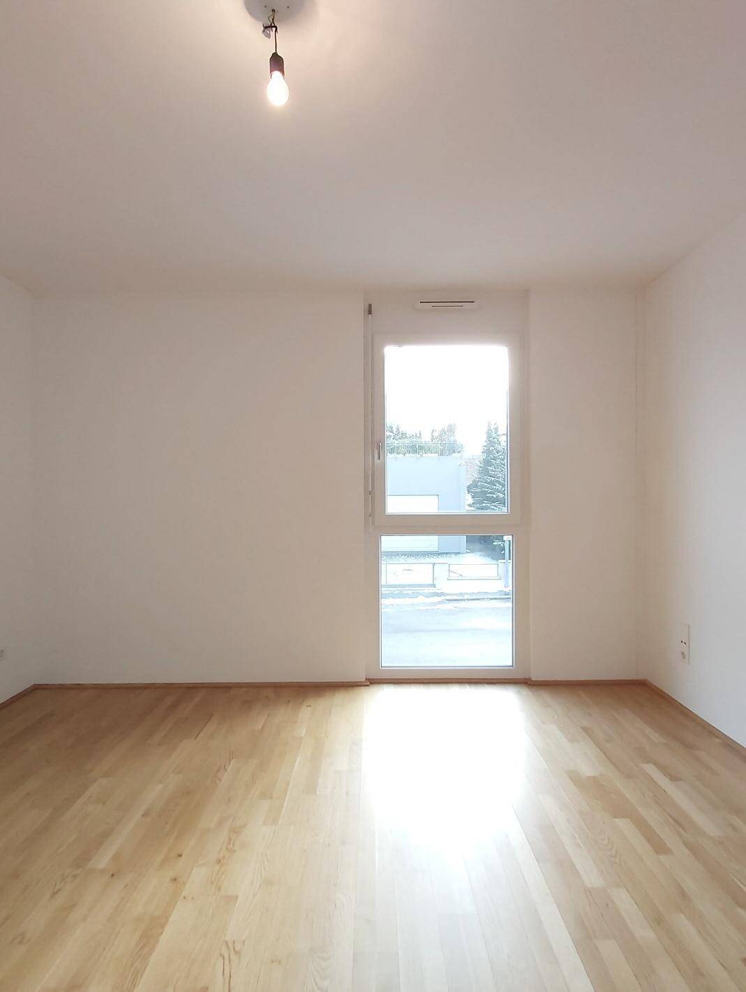 Helles Zimmer mit Parkettboden und einem hohen Fenster, das viel Tageslicht hereinlässt.