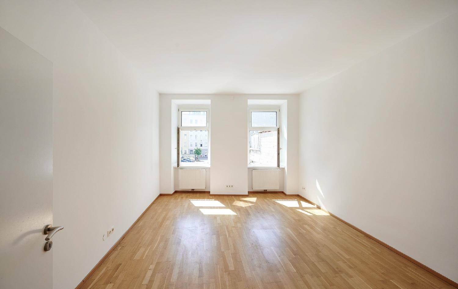 Heller, leerer Raum mit Parkettboden und zwei Fenstern, die viel Tageslicht hereinlassen.