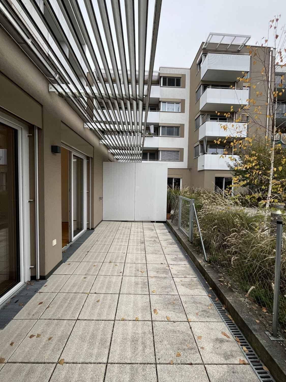 Große, geflieste Terrasse mit Pergola-Struktur und Blick auf die Wohnanlage.