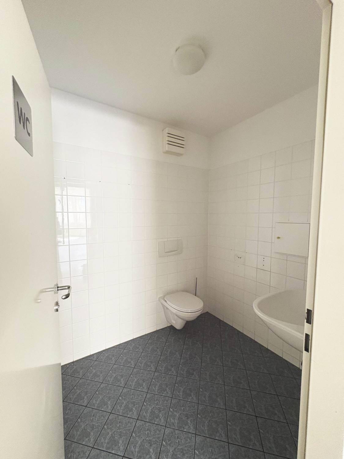 Saubere Toilette mit weißen Wandfliesen, einem Waschbecken und dunklem Fliesenboden.