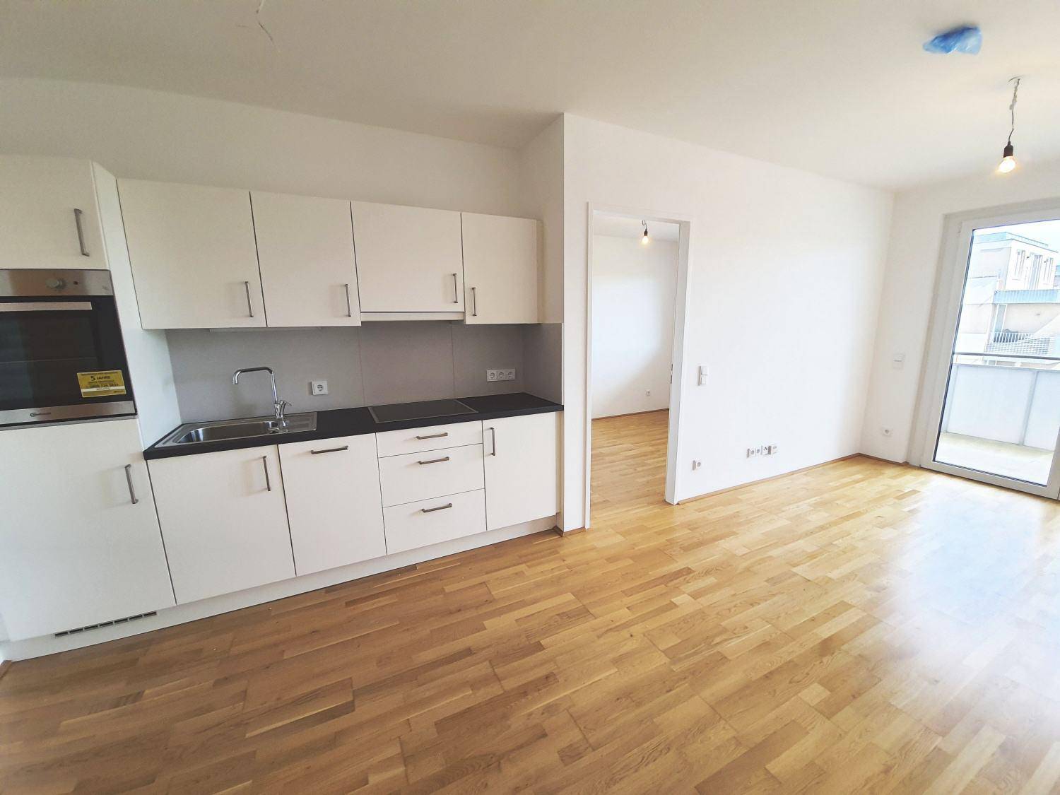 Heller offener Wohnbereich mit integrierter Küchenzeile, Parkettboden und Zugang zum Balkon.