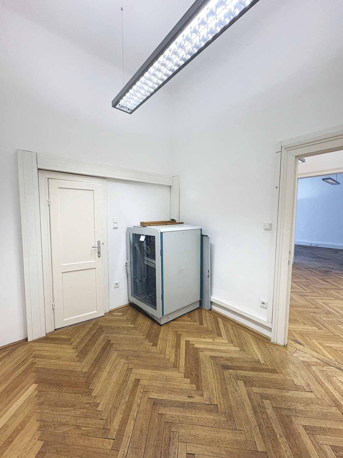 Ecke eines Zimmers mit Fischgrätparkett und einem Schrank, möglicherweise für Technik oder Lagerung.
