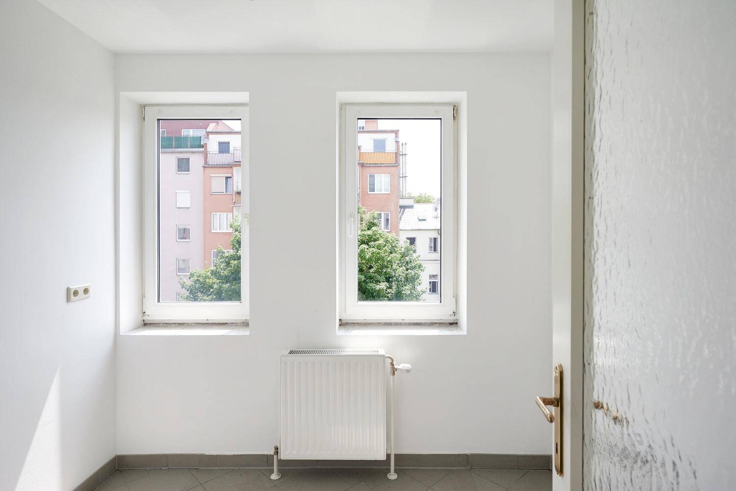 Kleiner Raum mit Fliesenboden, zwei Fenstern und Blick auf umliegende Gebäude.