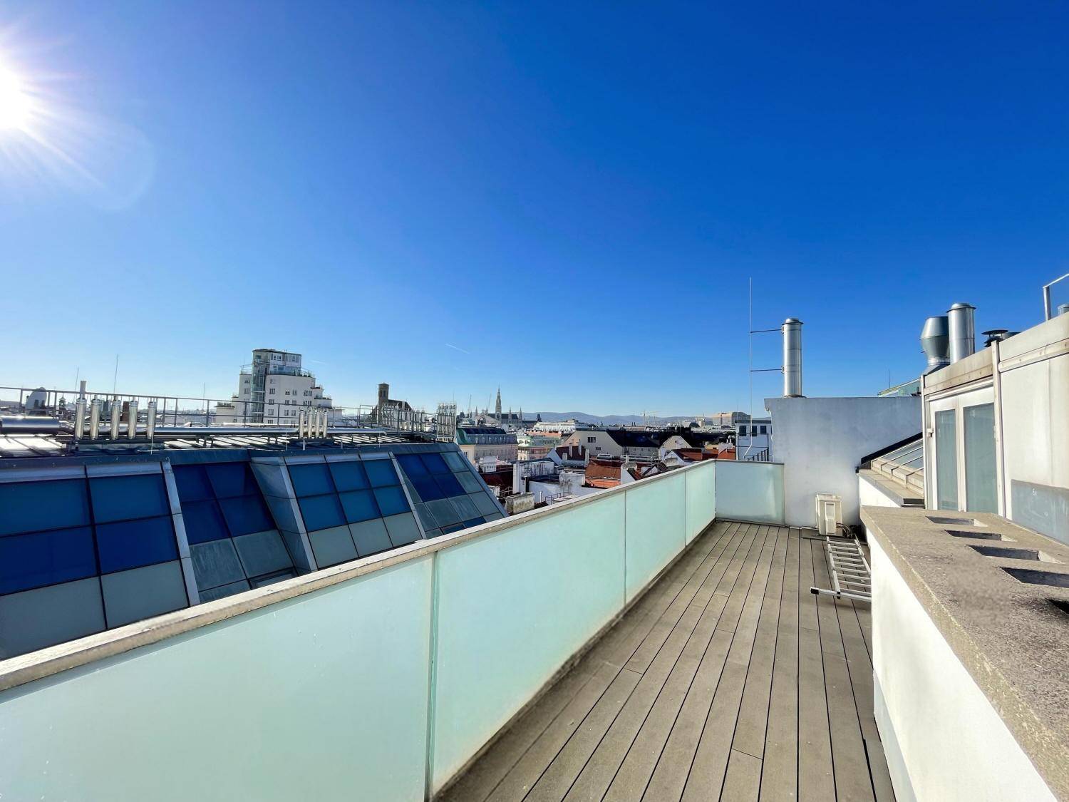 Sonniger Dachterrassenbereich mit Glasgeländer und weitem Blick über die Dächer der Stadt.