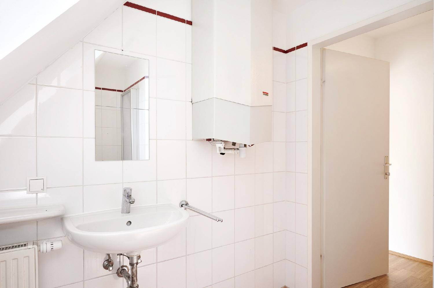 Badezimmer mit weißen Fliesen, Waschbecken und einem Warmwasserbereiter an der Wand.