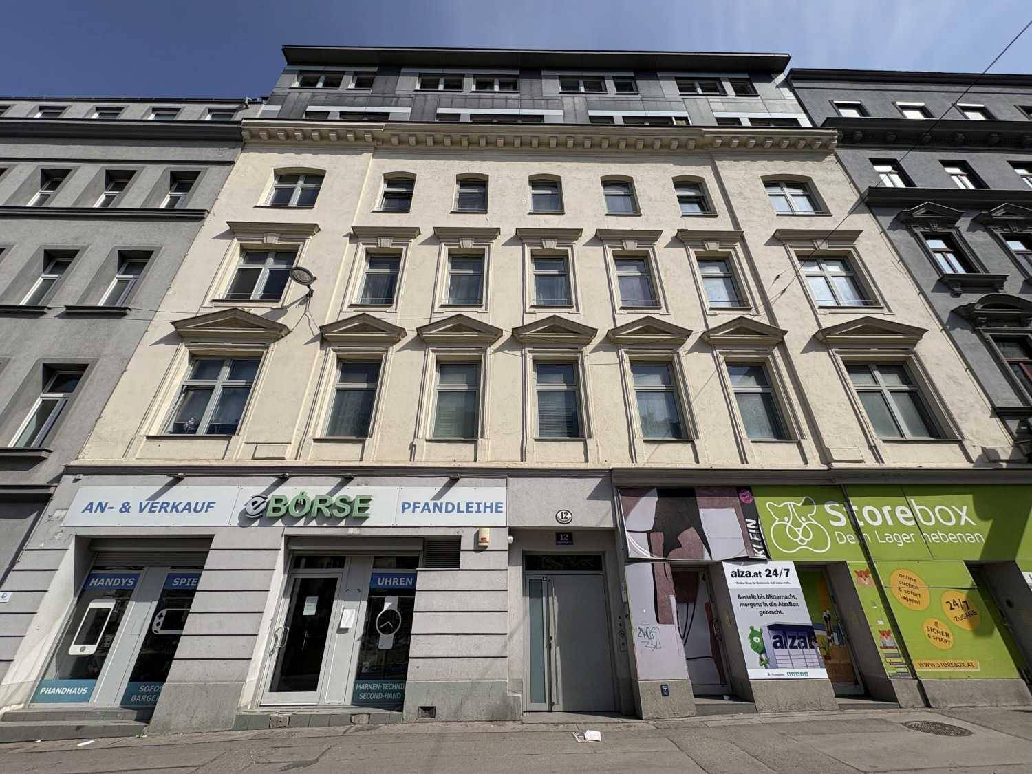 Historische Fassade eines Mehrfamilienhauses mit Geschäften im Erdgeschoss in einer städtischen Umgebung.
