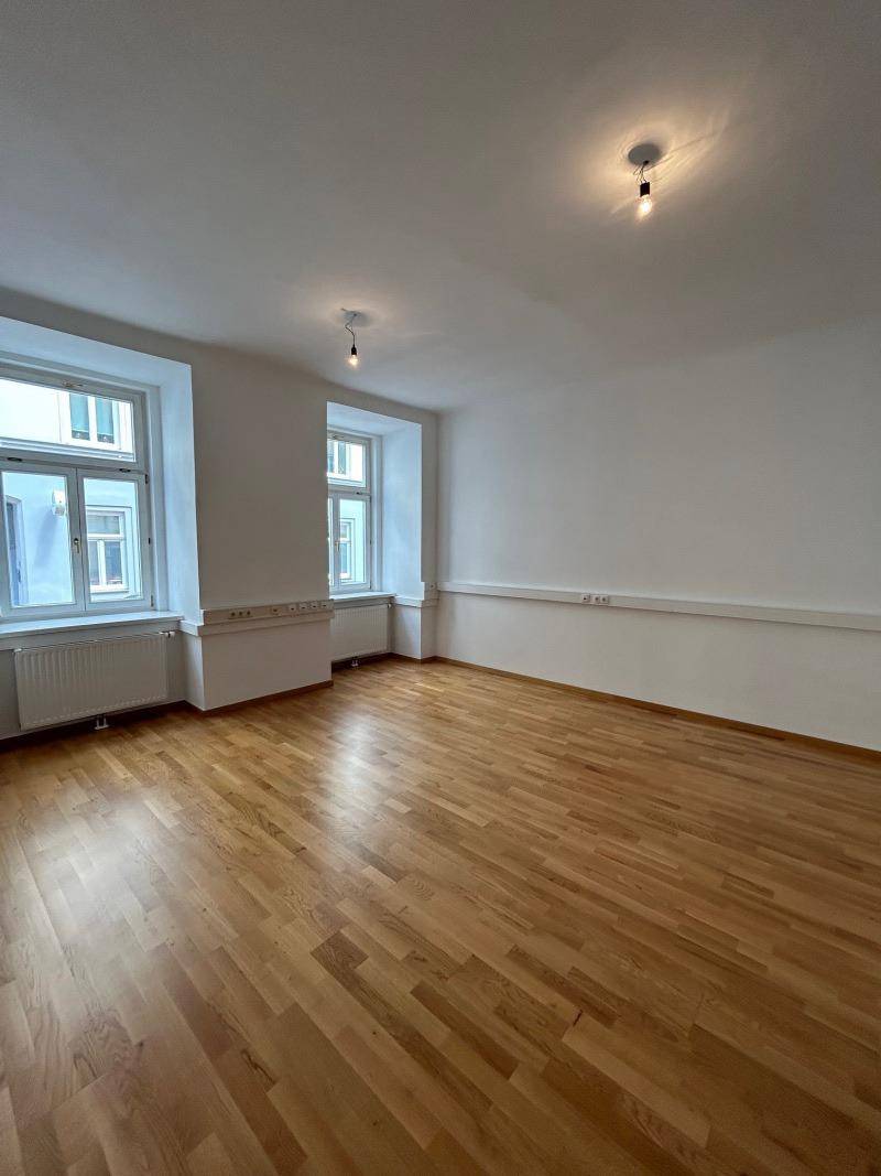 Geräumiges Zimmer mit zwei Fenstern, Holzboden und weißen Wänden.