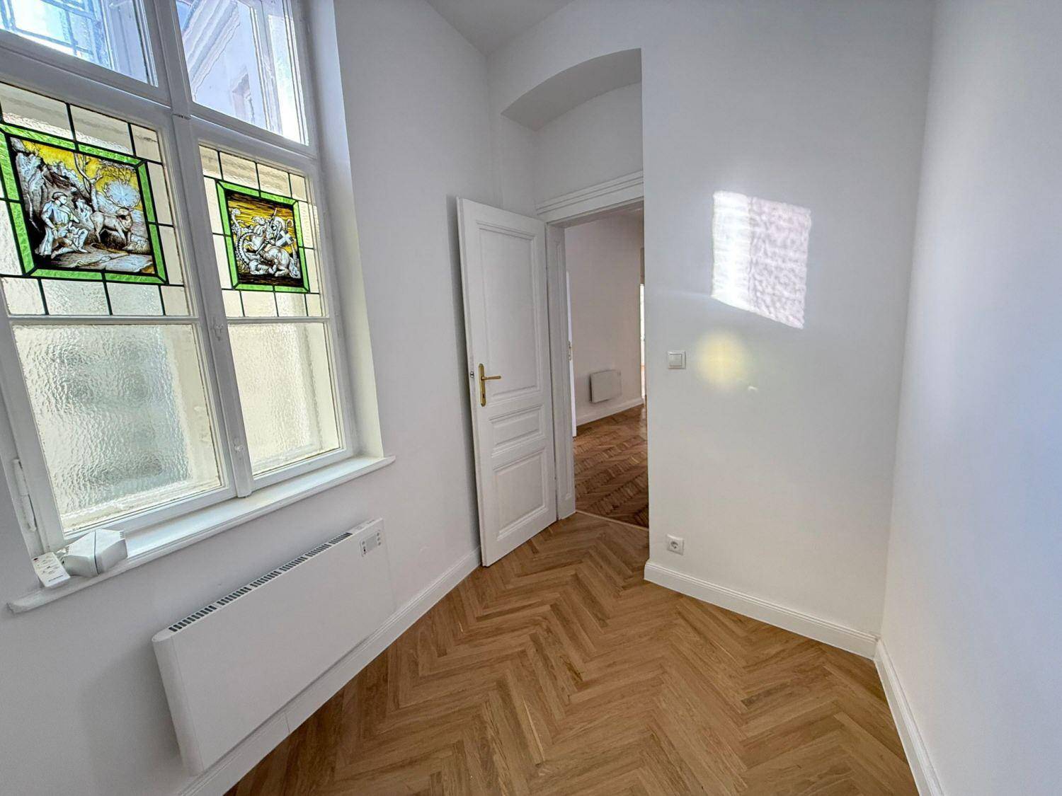Heller Raum mit Fischgrätparkett, Heizkörper und einem Fenster mit dekorativen Buntglaselementen.