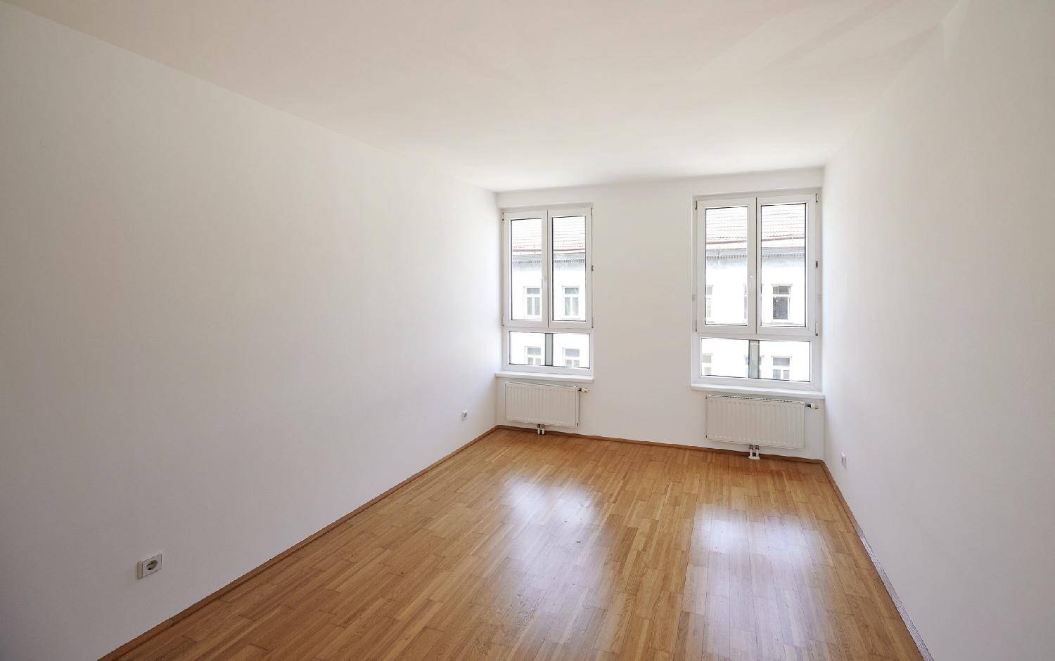 Helles Zimmer mit Holzboden, weißen Wänden und zwei Fenstern mit Blick auf die Straße.