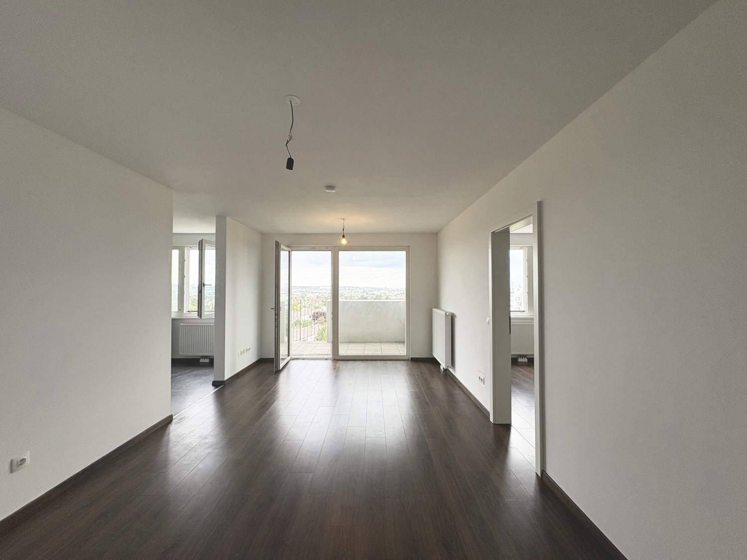 Helles Wohnzimmer mit dunklem Laminatboden, weißen Wänden und Zugang zum Balkon mit Weitblick.