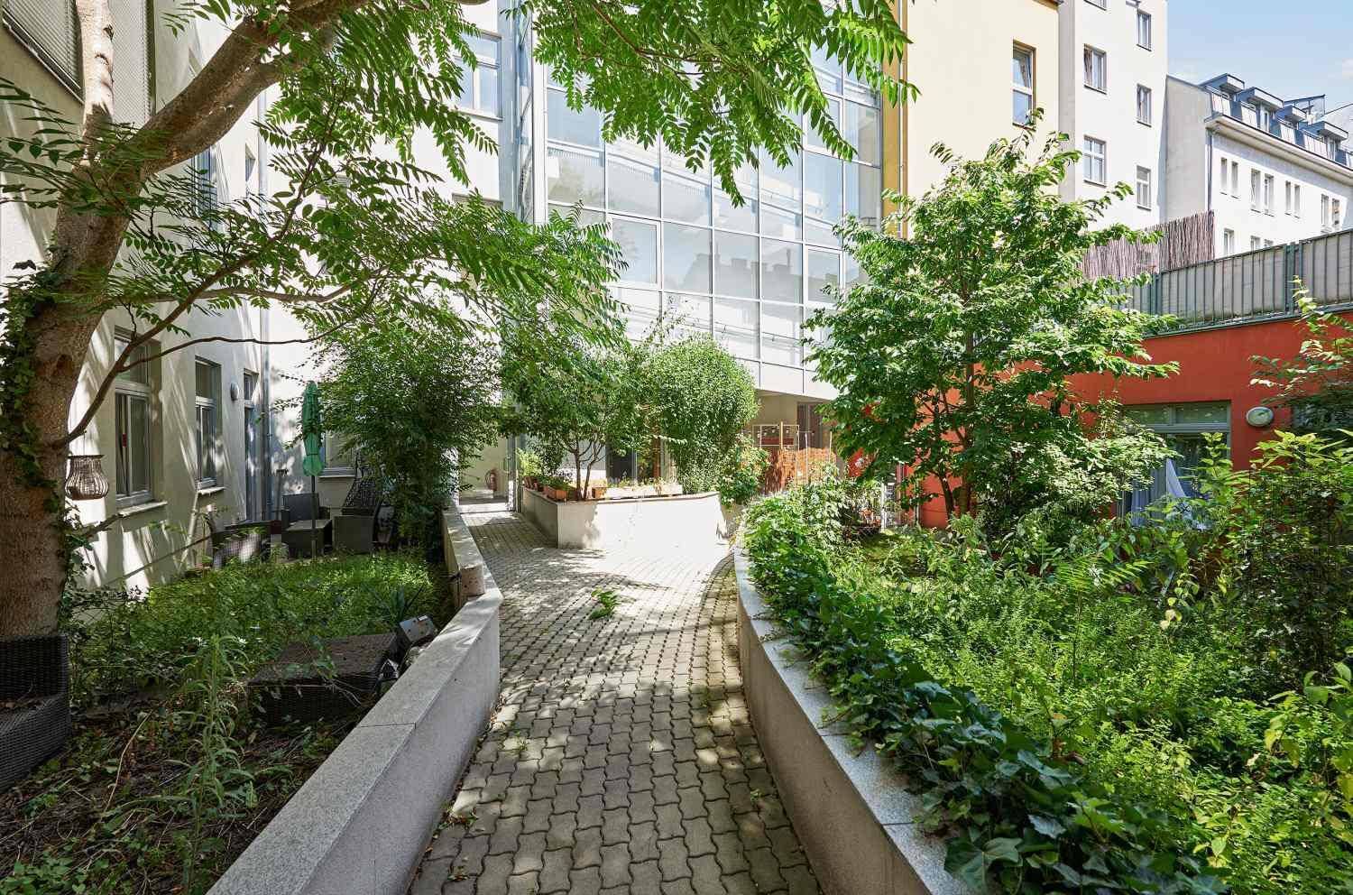 Sonniger Innenhof mit üppiger Bepflanzung, einem gepflasterten Weg und modernen Gebäuden.