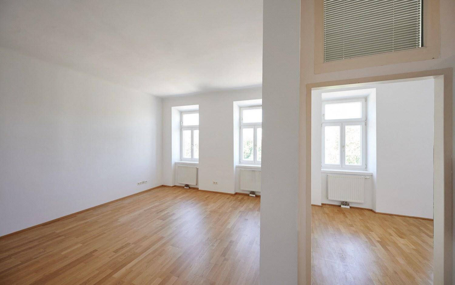 Geräumiges, helles Zimmer mit Parkettboden und Blick in einen weiteren Raum.