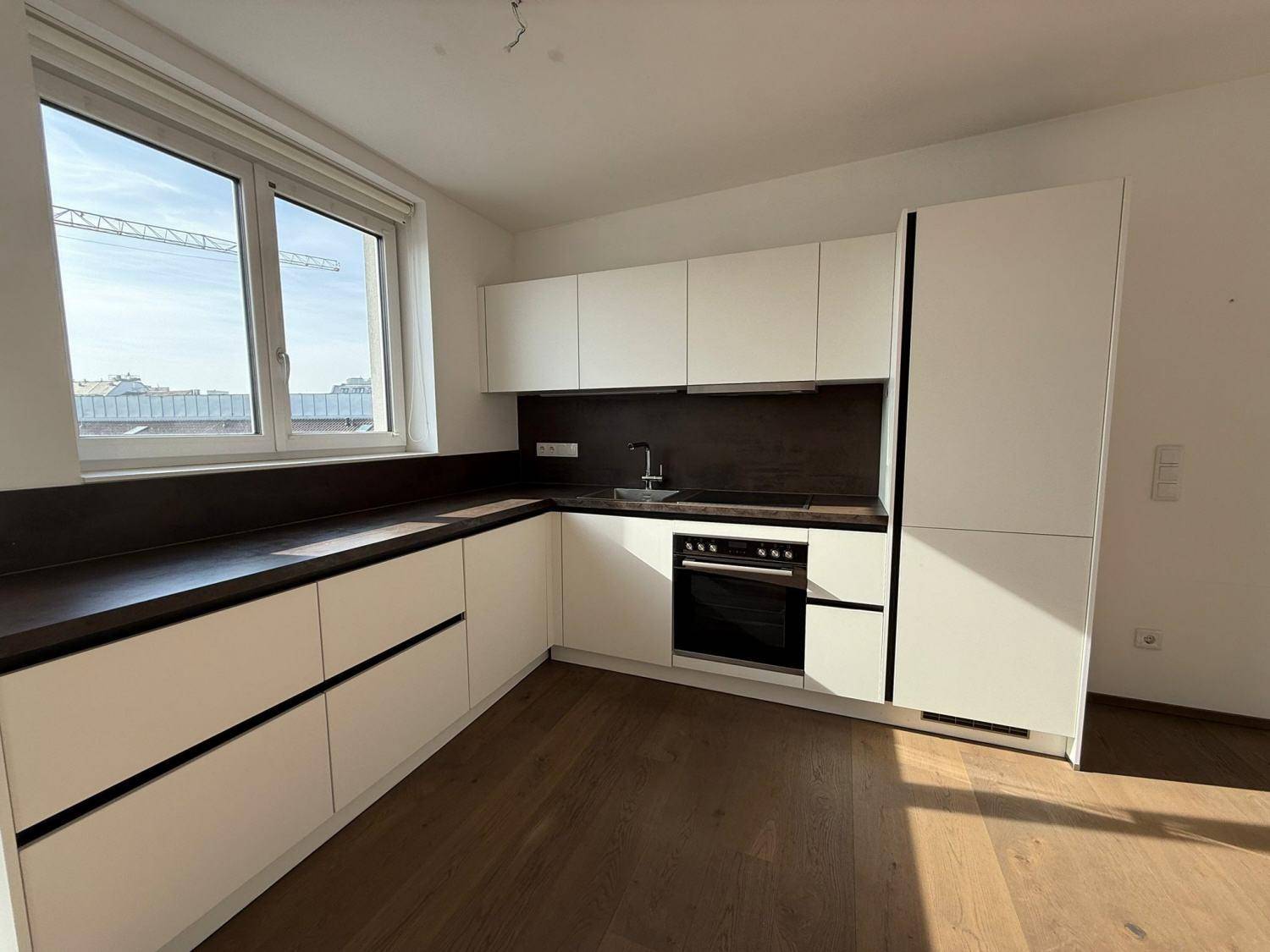 Moderne weiße Einbauküche mit dunkler Arbeitsplatte und integriertem Backofen unter einem Fenster.
