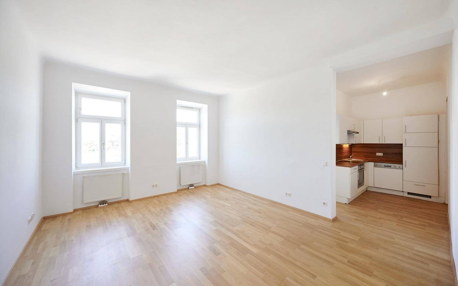 Helles, leeres Zimmer mit Parkettboden und zwei Fenstern, offen zur Küche.