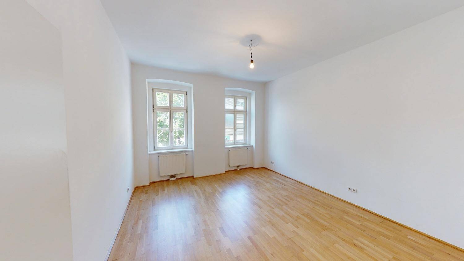 Leeres Schlafzimmer mit Parkettboden, weißen Wänden und zwei klassischen Fenstern.
