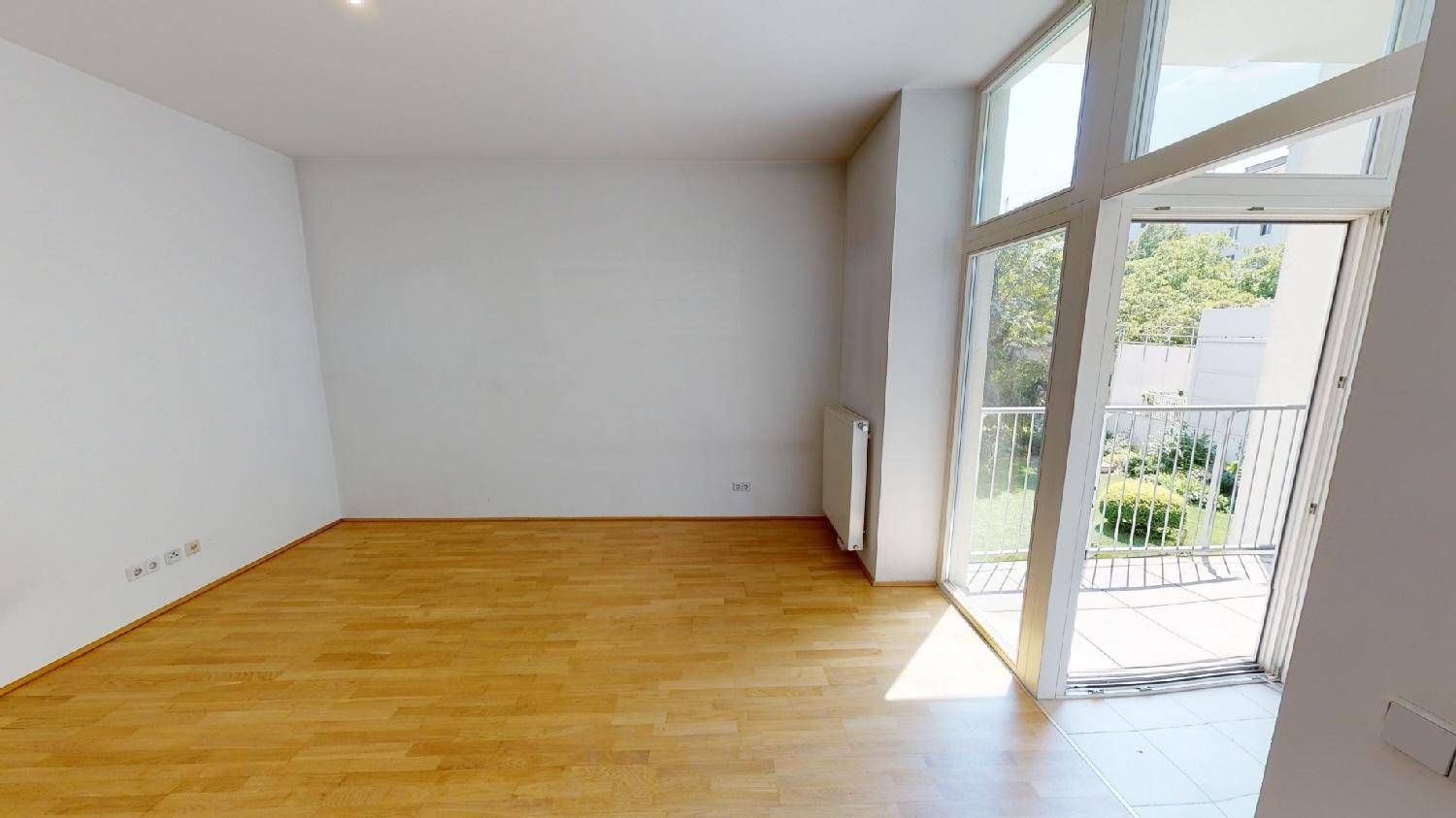 Helles, leeres Wohnzimmer mit Parkettboden und Zugang zu einem Balkon.