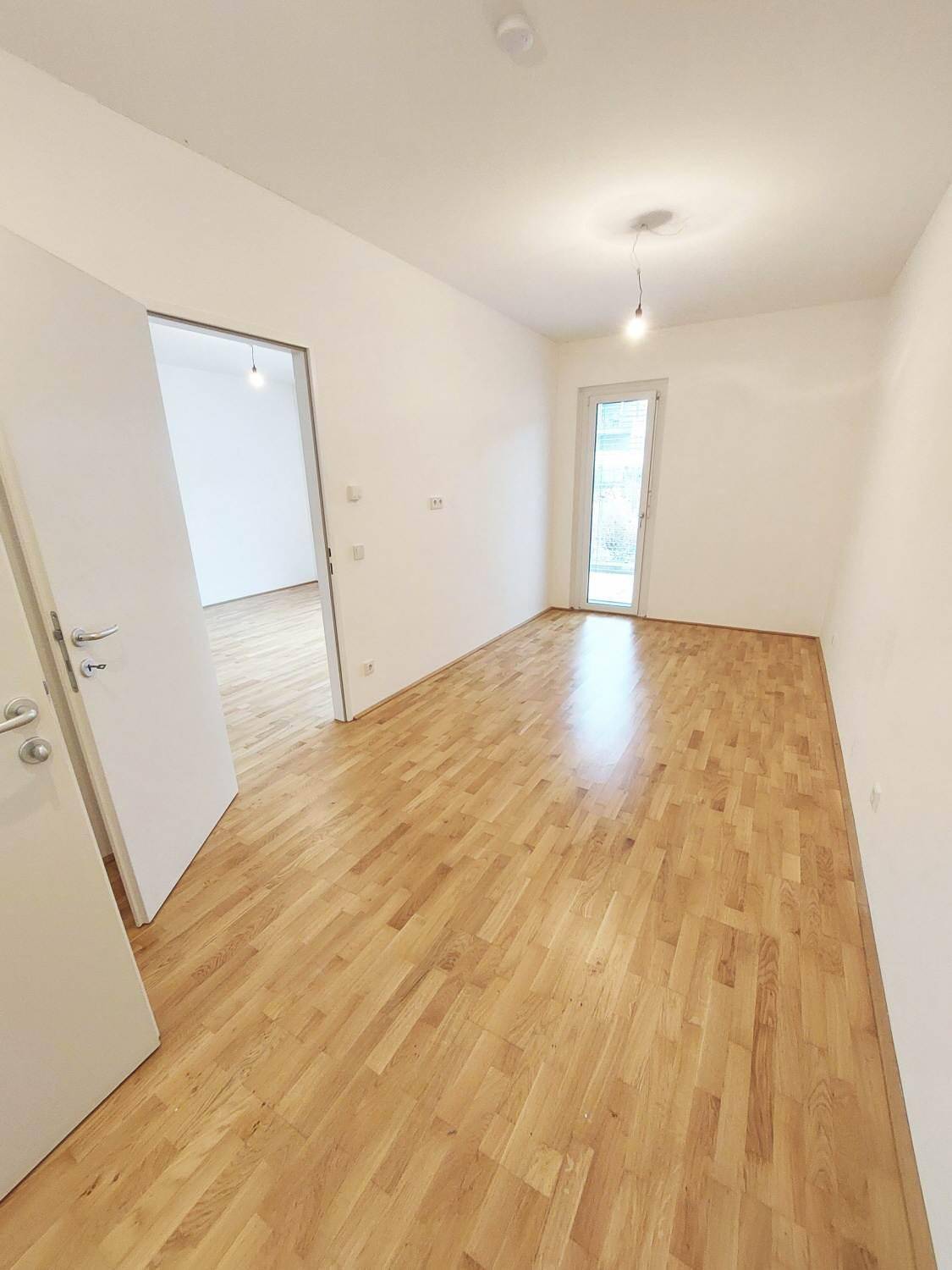 Geräumiges Zimmer mit Parkettboden, weißen Wänden und Zugang zu einem Außenbereich.