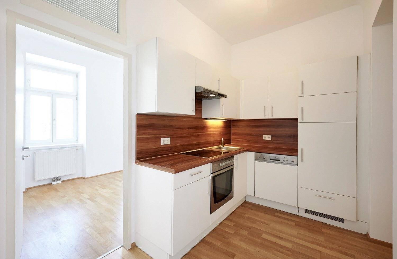 Kompakte Einbauküche mit weißen Fronten, Holzarbeitsplatte und Blick in ein Nebenzimmer.
