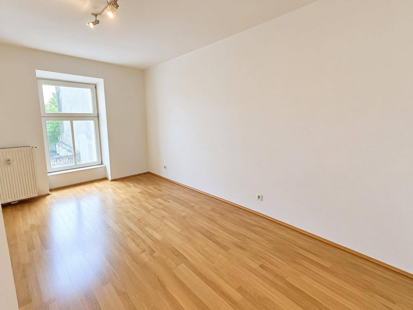 Leeres Zimmer mit Parkettboden und einem großen Fenster, das den Raum mit natürlichem Licht füllt.