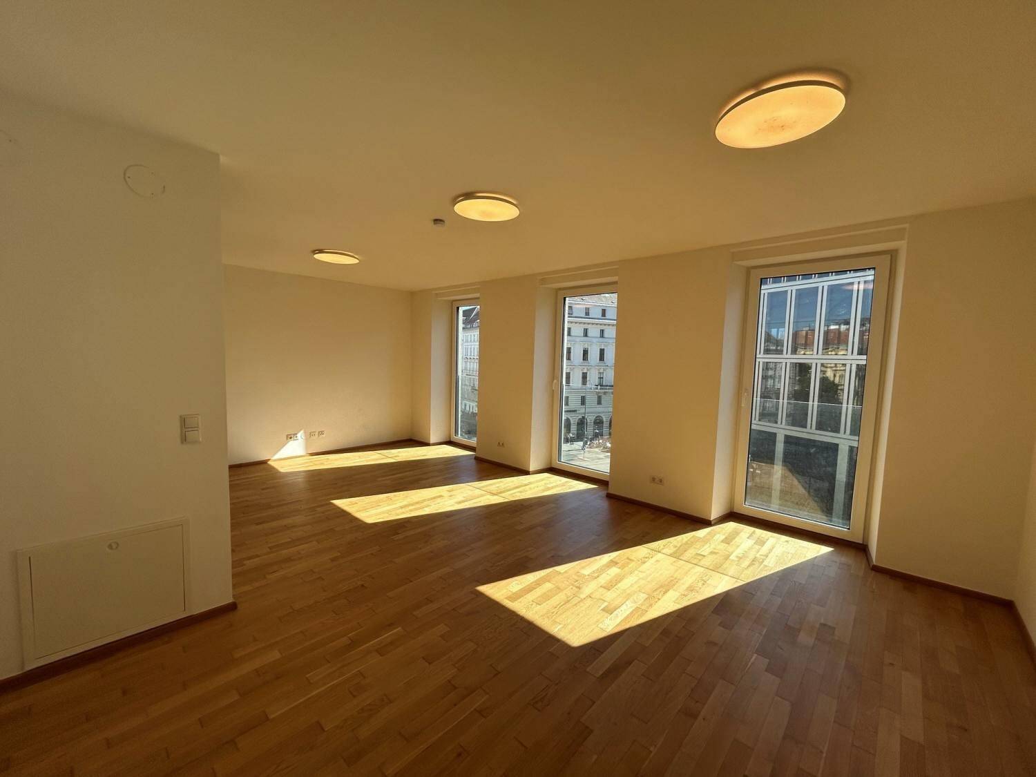 Helles, leeres Wohnzimmer mit Holzboden und großen Fenstern, durch die Sonnenlicht fällt.
