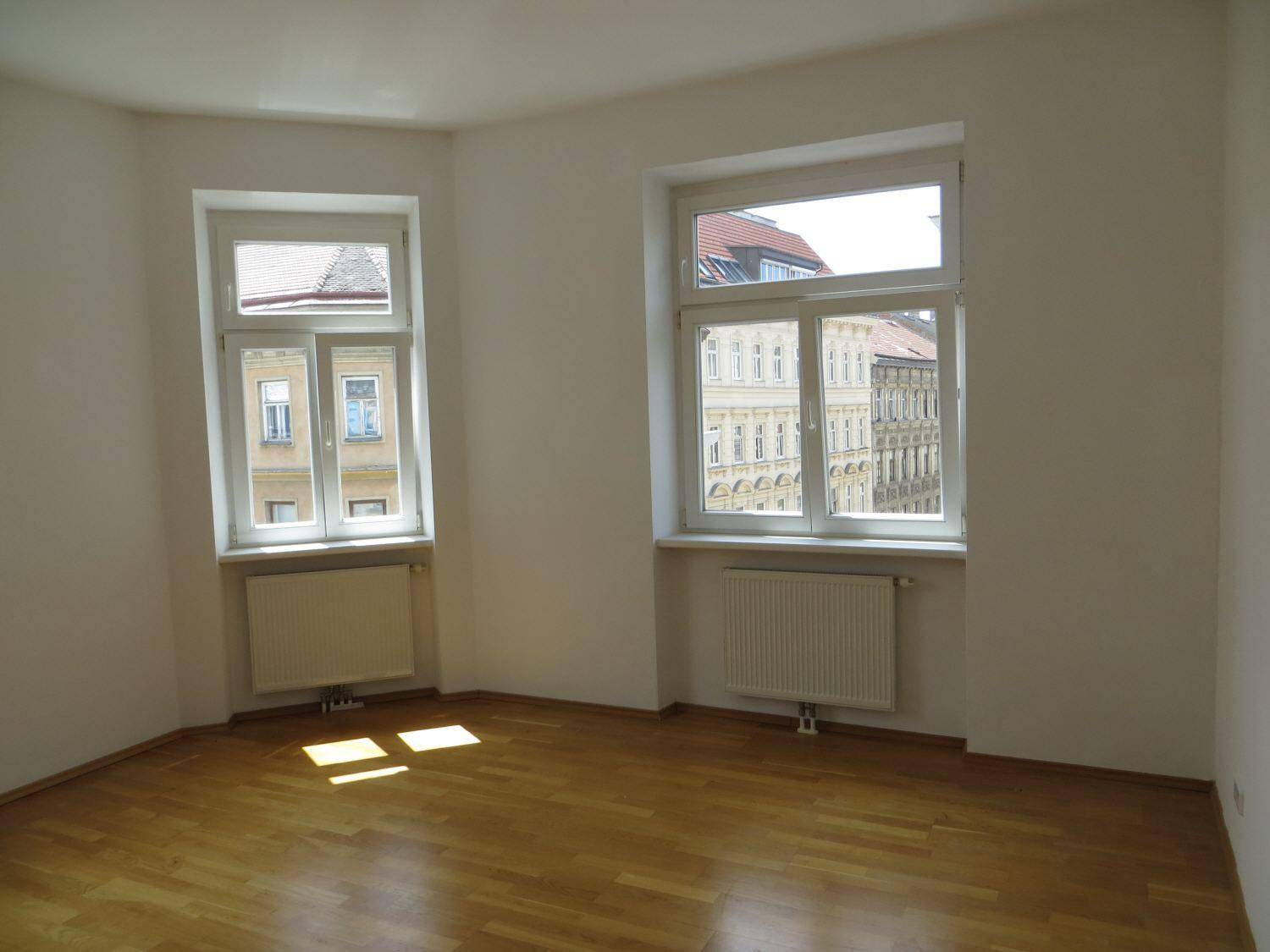 Helles Zimmer mit Parkettboden und zwei Fenstern, die viel Tageslicht hereinlassen.