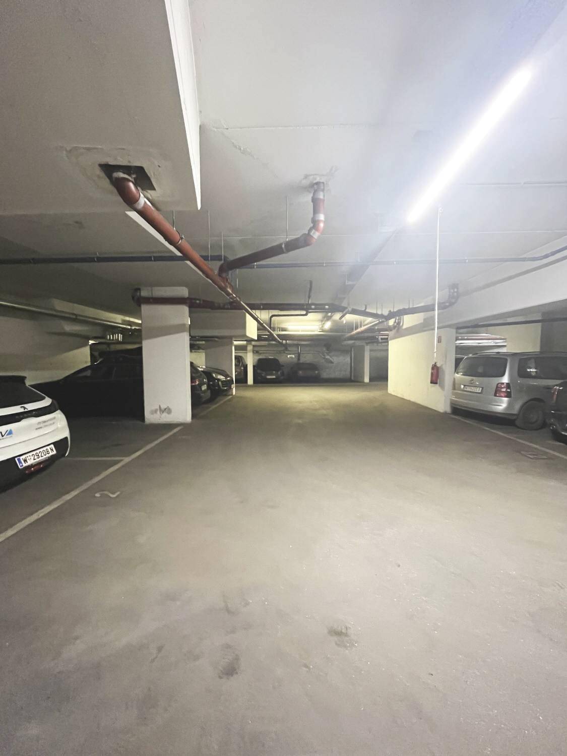 Weitläufige Tiefgarage mit sichtbaren Rohren an der Decke und mehreren geparkten Autos.