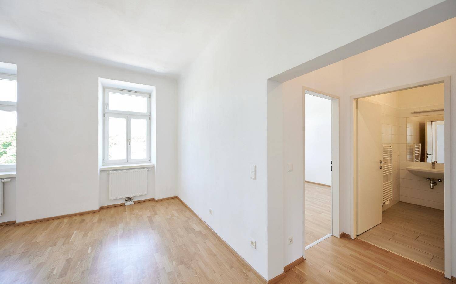 Heller Raum mit Parkettboden und Blick in ein gefliestes Badezimmer mit Waschbecken.
