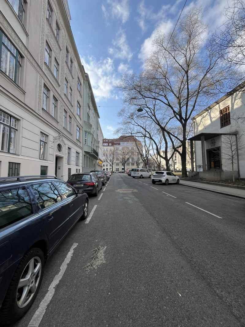 Blick die Straße entlang mit parkenden Autos, Bäumen und weiteren Gebäuden unter blauem Himmel.