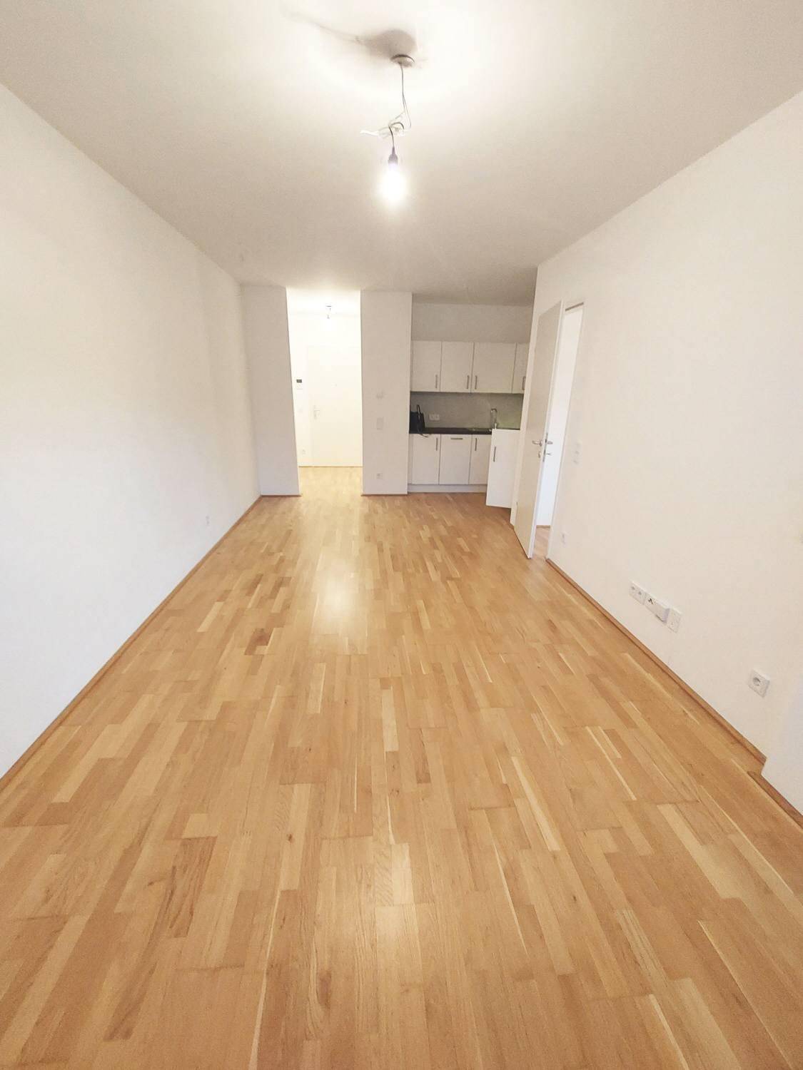 Geräumiger Wohnbereich mit Parkettboden, weißer Küchenzeile im Hintergrund und Balkonzugang.