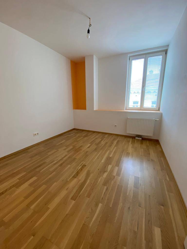 Helles Zimmer mit Parkettboden und einem Fenster, eine orangefarbene Nische setzt Akzente.