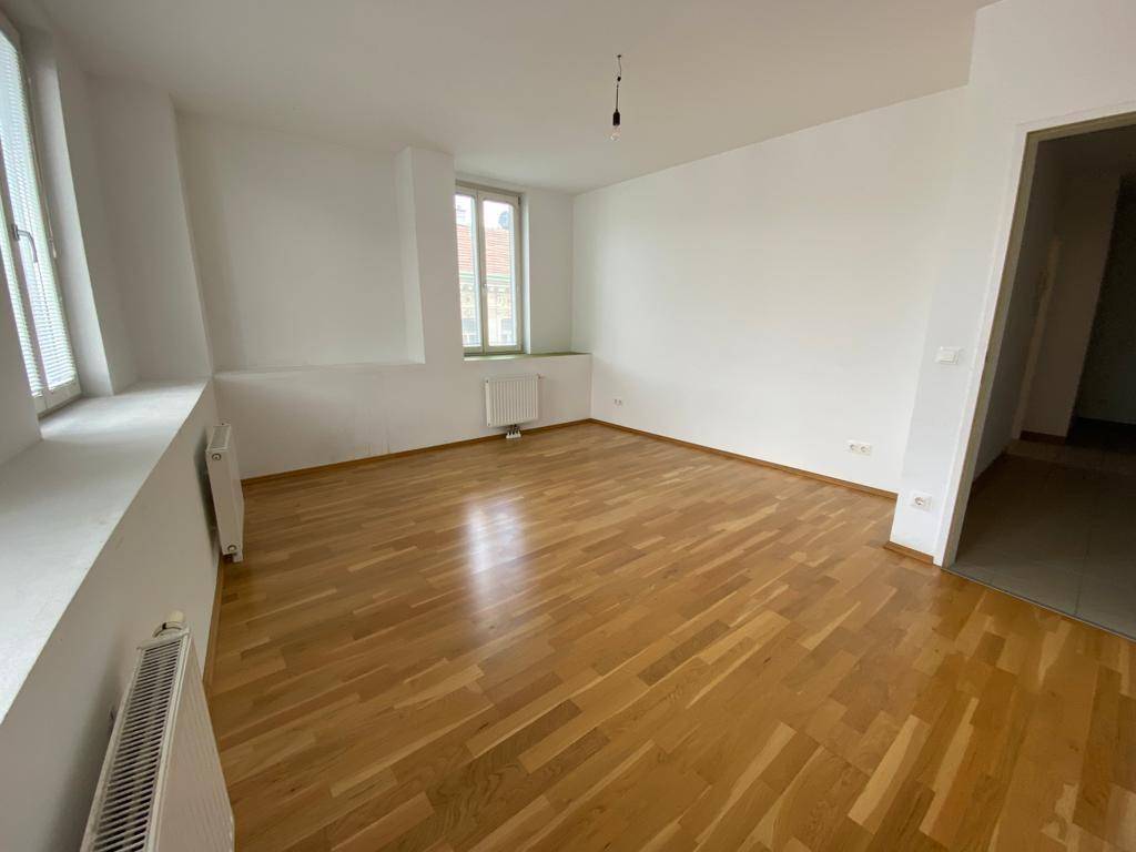 Helles, leeres Zimmer mit Parkettboden und zwei Fenstern, ideal für ein Wohnzimmer oder Büro.