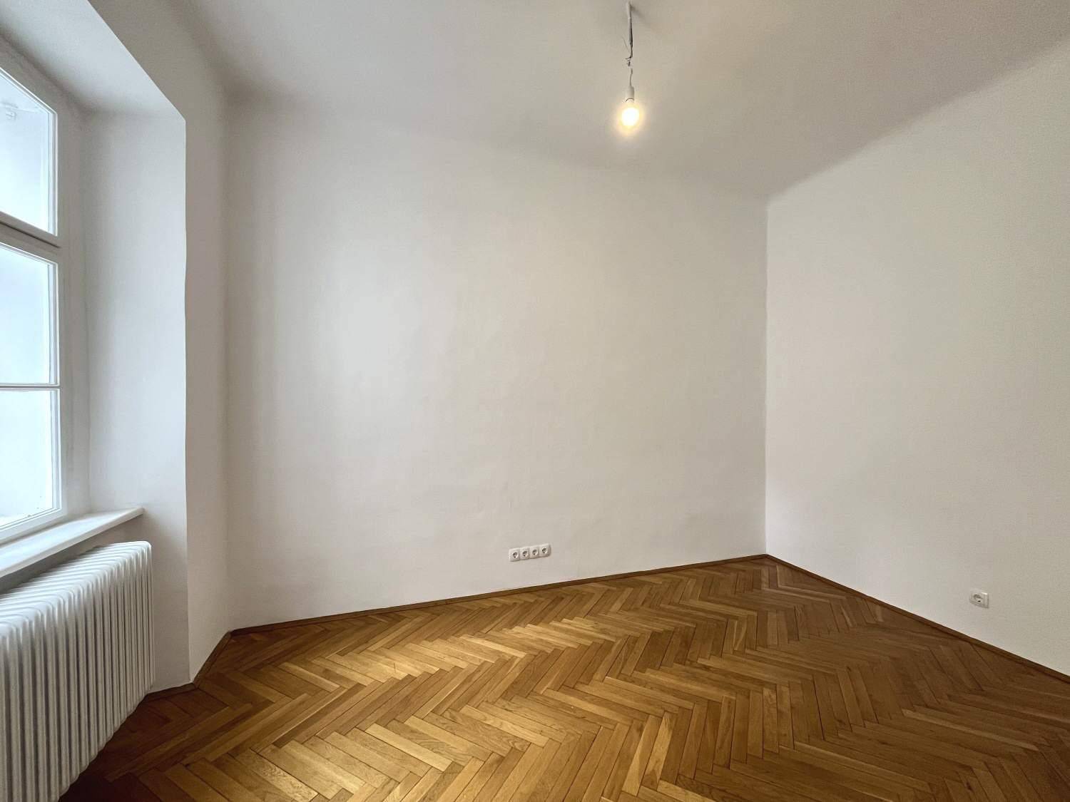 Helles Zimmer mit Fischgrätparkett und einem großen Fenster, das viel Tageslicht hereinlässt.