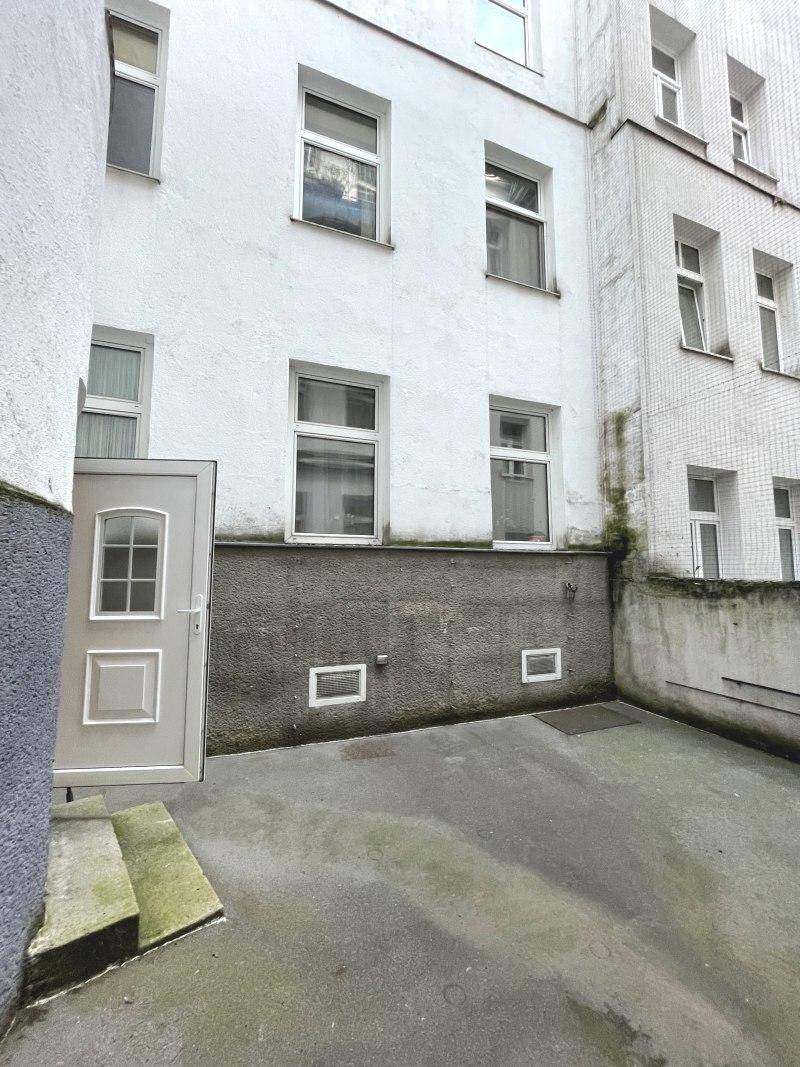Kleiner, gepflasterter Bereich im Hinterhof mit einer Tür und einer grauen Wand.