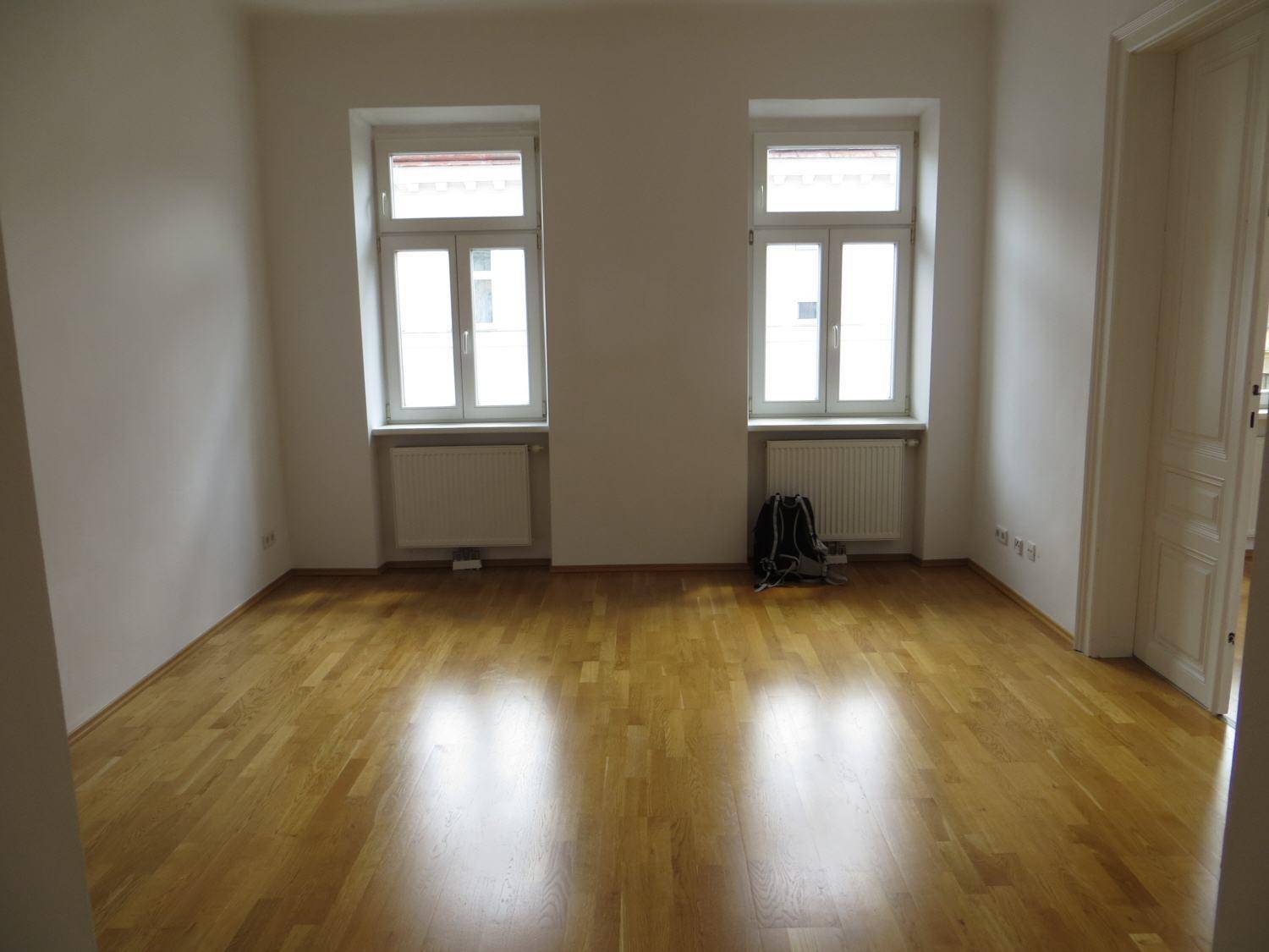 Helles, leeres Zimmer mit Parkettboden und zwei Fenstern, bereit zur individuellen Gestaltung.