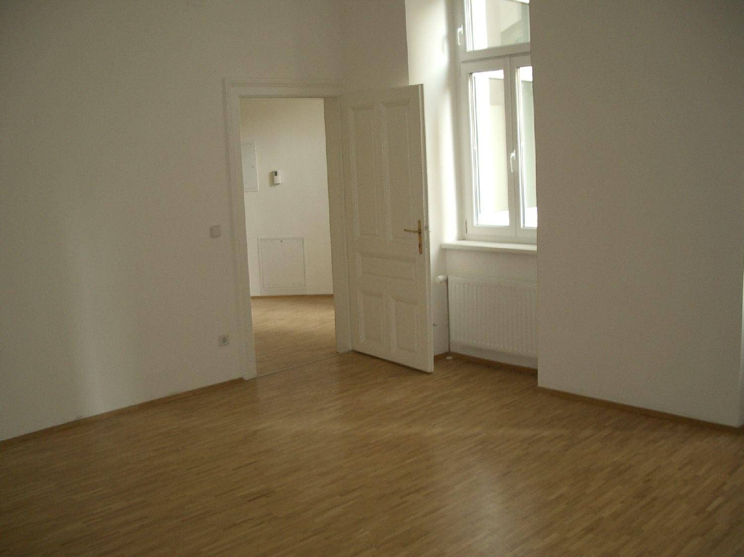Leerer Raum mit hellem Parkettboden, einem Fenster und einer offenen Tür zu einem weiteren Bereich.