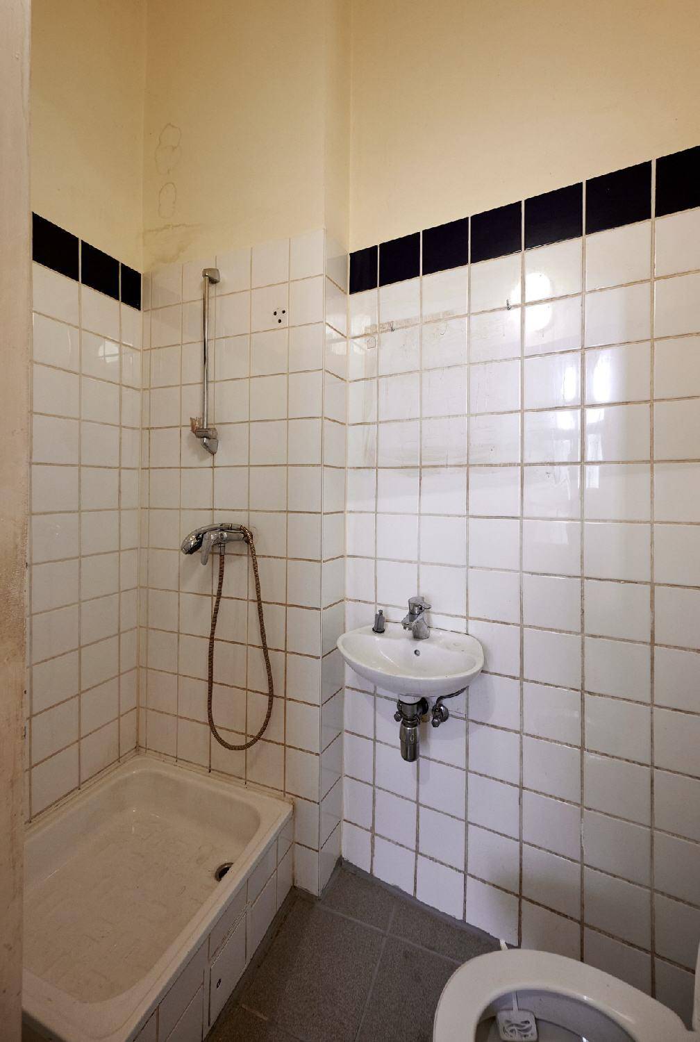 Kleines Badezimmer mit weißen Fliesen, Duschwanne, Waschbecken und Toilette, renovierungsbedürftig.