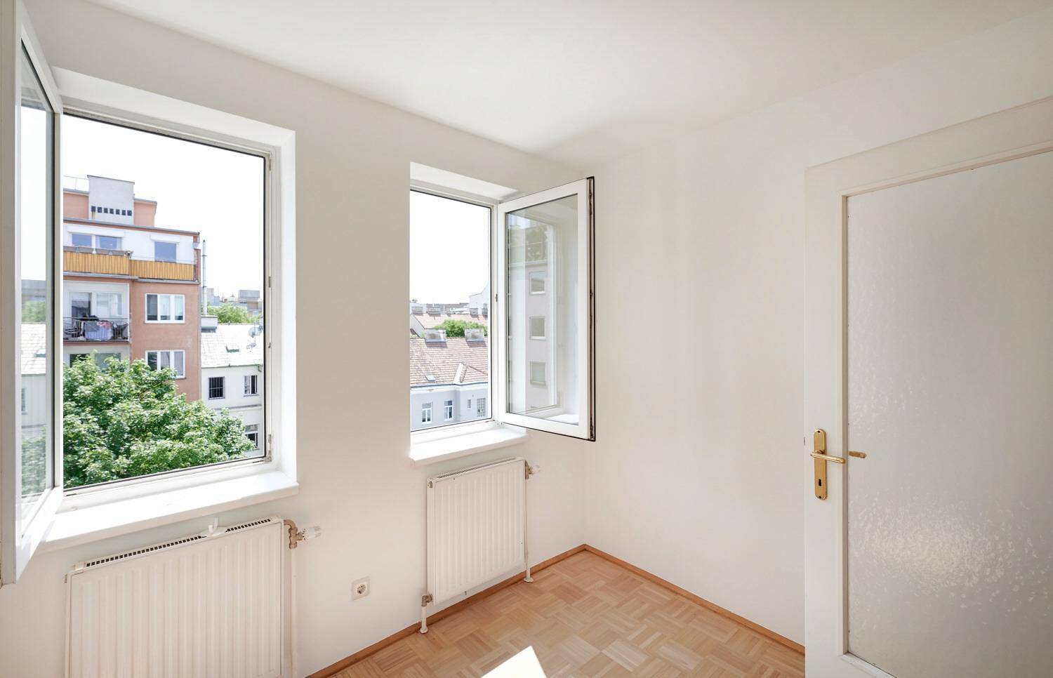 Helles Zimmer mit Parkettboden und zwei geöffneten Fenstern, die einen Blick auf die Stadt bieten.